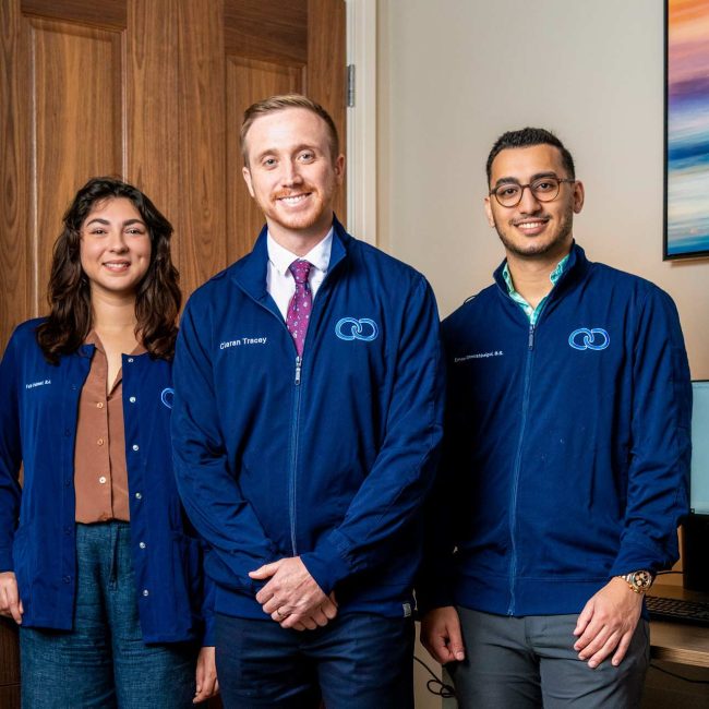 Three team members in navy Caron-branded jackets standing together in an office, featured in a Boca Observer profile.