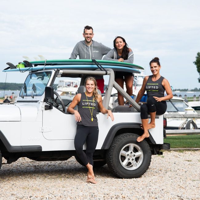 Fitness team wearing LIFTED-branded activewear, posed on and around a Jeep with paddleboards near a marina.
