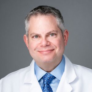 Headshot of a man wearing a white lab coat over a light blue shirt and patterned tie.