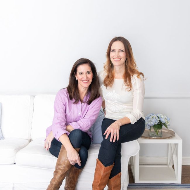 Two women sitting together on a white couch in a bright, casual setting.