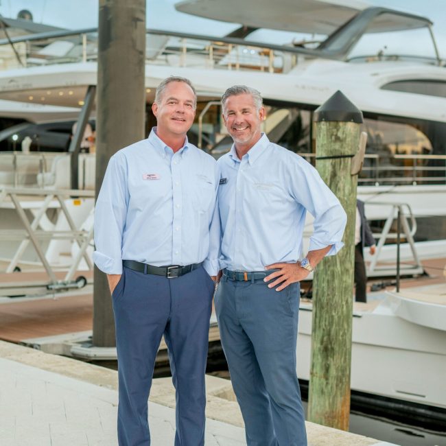 Two yacht executives standing together at a marina, wearing blue shirts and navy pants, with a yacht docked behind them.