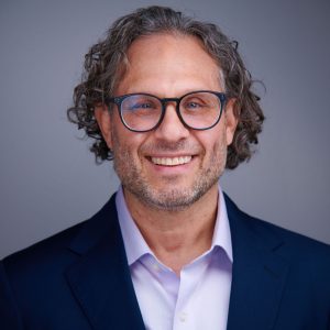 Headshot of a smiling man with curly hair and glasses, wearing a navy blazer.