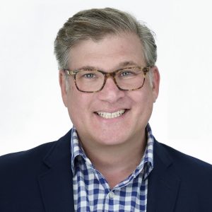 Headshot of a man wearing tortoiseshell glasses, a navy blazer, and a blue checked shirt.