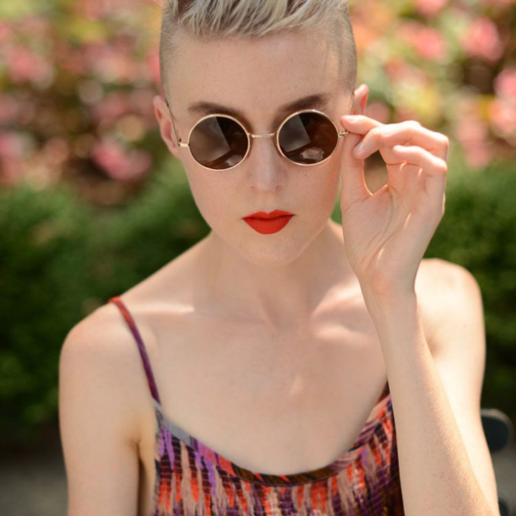 Woman with short blonde hair and round sunglasses, wearing a colorful dress