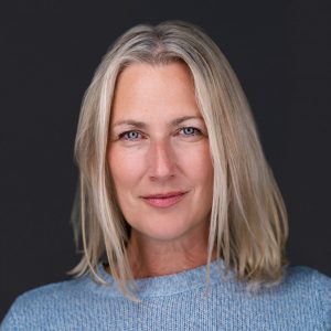 Headshot of a woman with blonde hair and a blue sweater against a dark background.