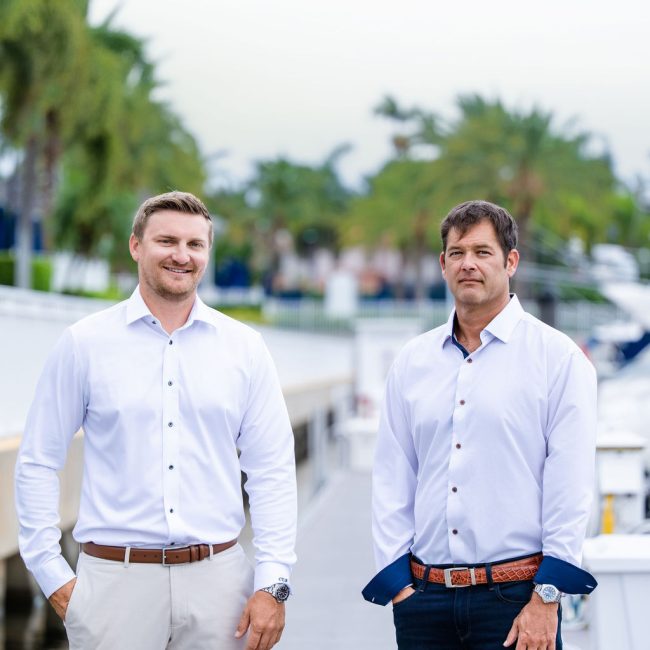 Two yacht brokers standing together at a marina with boats in the background.